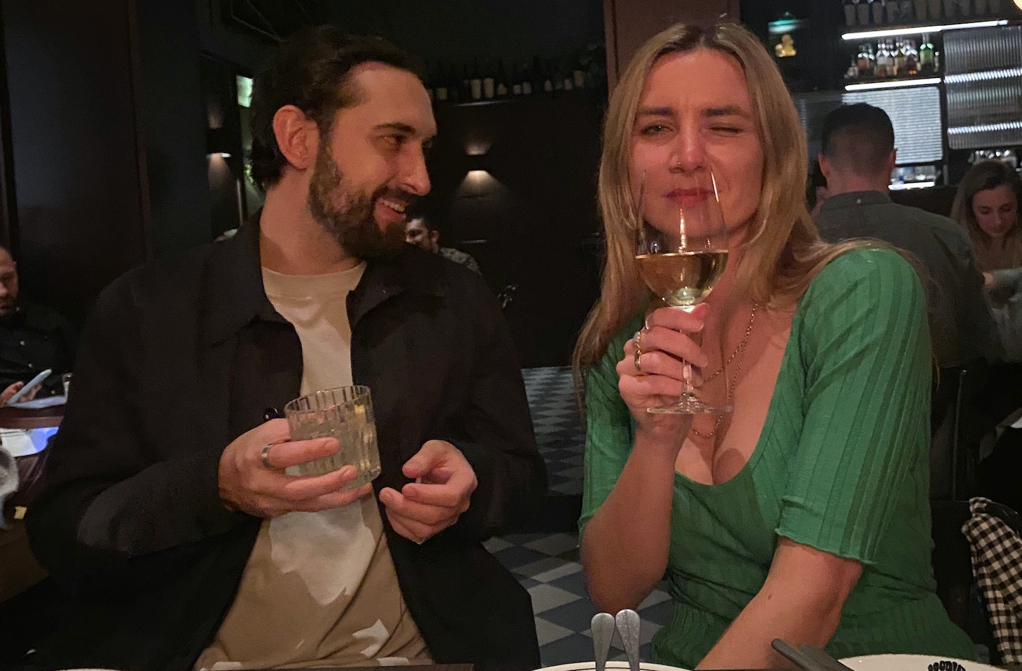 A man and woman sitting in a moody restaurant hold their drinks. The woman winks at the camera.